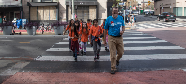 Students in orange shirts cross the street with an adult tour guide.