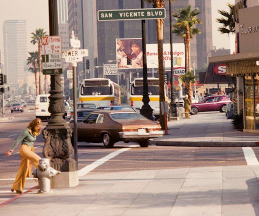 Image of Beverly Hills in the 1970s with period cars turning a busy street and a young woman crossing the street.