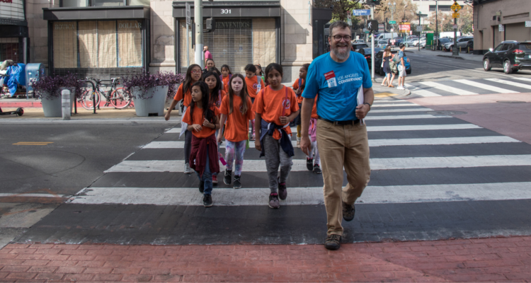 Students in orange shirts cross the street with an adult tour guide.