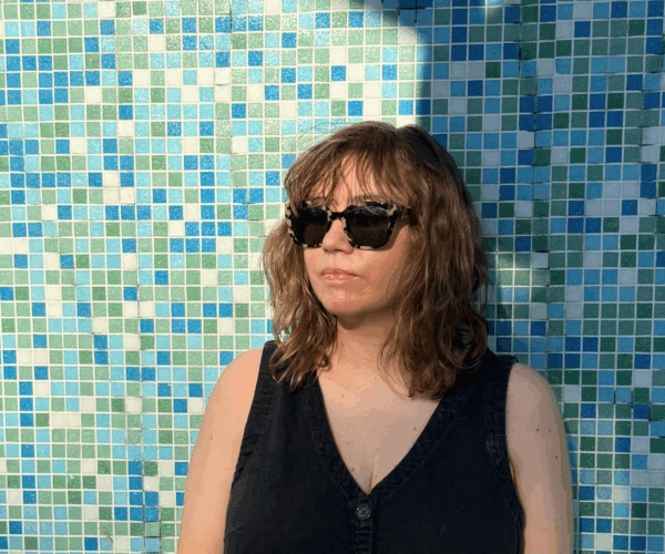 Woman wearing sunglasses stands in front of a blue, green, teal, and white tiled wall.