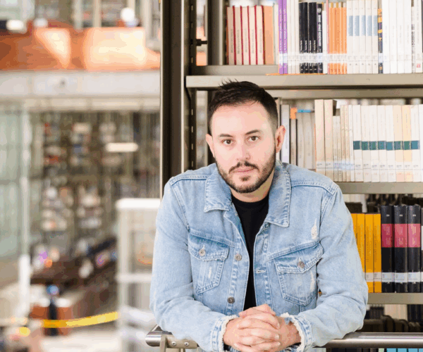 A man wearing a jean jacket with a black undershirt stand in front of a bookshelf with a scenic library behind him.