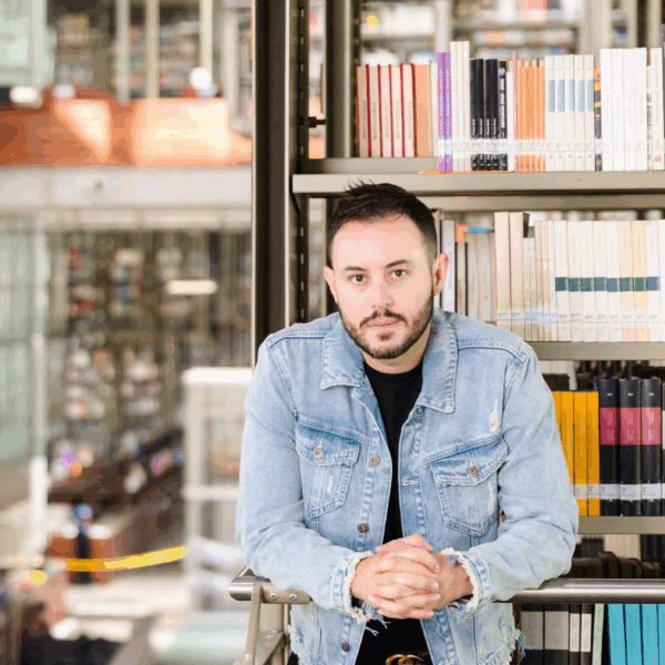 A man wearing a jean jacket with a black undershirt stand in front of a bookshelf with a scenic library behind him.