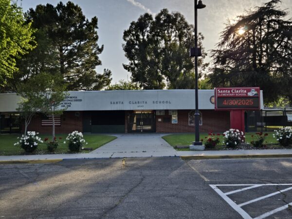 single story brick clad school building visible from asphalt parking lot with large trees peaking over the flat roof from the center courtyard.