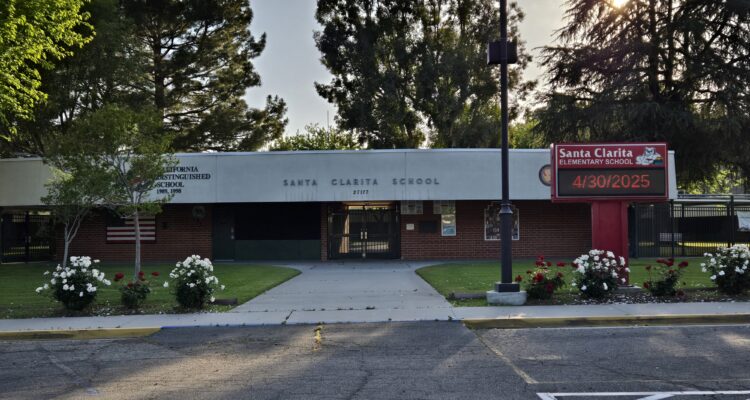 single story brick clad school building visible from asphalt parking lot with large trees peaking over the flat roof from the center courtyard.