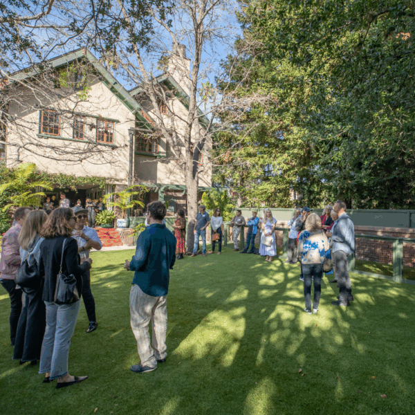 A group of people of various ages standing in a grassy yard, engaged in conversation in a sunny environment.