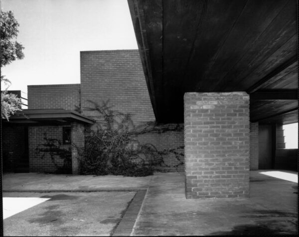 Photograph of an exterior view of the Sturges House, Los Angeles. An inner courtyard is shown in the image. In the foreground, on top, a roof extends toward the middle of the image partially covering the courtyard. In the middle, at right, a brick pillar can be seen. In the background, a brick wall is visible, partially covered by vegetation. The structure was designed by architect Frank Lloyd Wright.