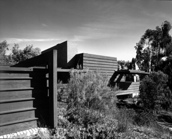 Photograph of an exterior view of the Sturges House, Los Angeles. In the foreground, at left, a wall constructed of wooden horizontal boards extends toward the middle of the image. In the middle, at right, a roof-covered deck can be partially seen. In the background, at center, a brick-constructed segment of the house is visible. The structure was designed by architect Frank Lloyd Wright.
