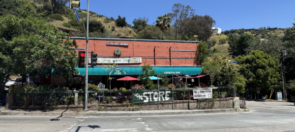 Masonry brick building with a smooth facade on the front primary elevation. The building includes a centered clock and canyon country store coca cola sign. There is a deck in front with ornamental plants surrounding the deck.