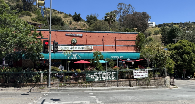 Masonry brick building with a smooth facade on the front primary elevation. The building includes a centered clock and canyon country store coca cola sign. There is a deck in front with ornamental plants surrounding the deck.
